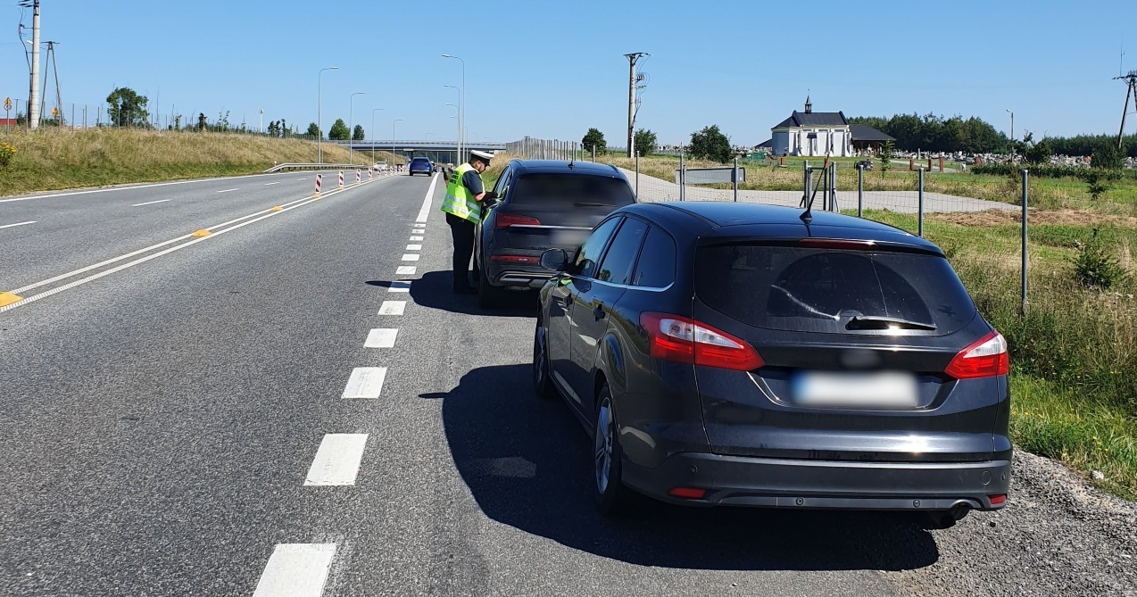 Na pierwszym planie: tył i lewy bok nieoznakowanego radiowozu Inspekcji Transportu Drogowego z zamontowanym fotorejestratorem prędkości. Przed nieoznakowanym pojazdem stoi samochód osobowy. Umundurowany inspektor Inspekcji Transportu Drogowego pobiera dokumenty od kierującego. W tle: droga ekspresowa i jadące po niej pojazdy.